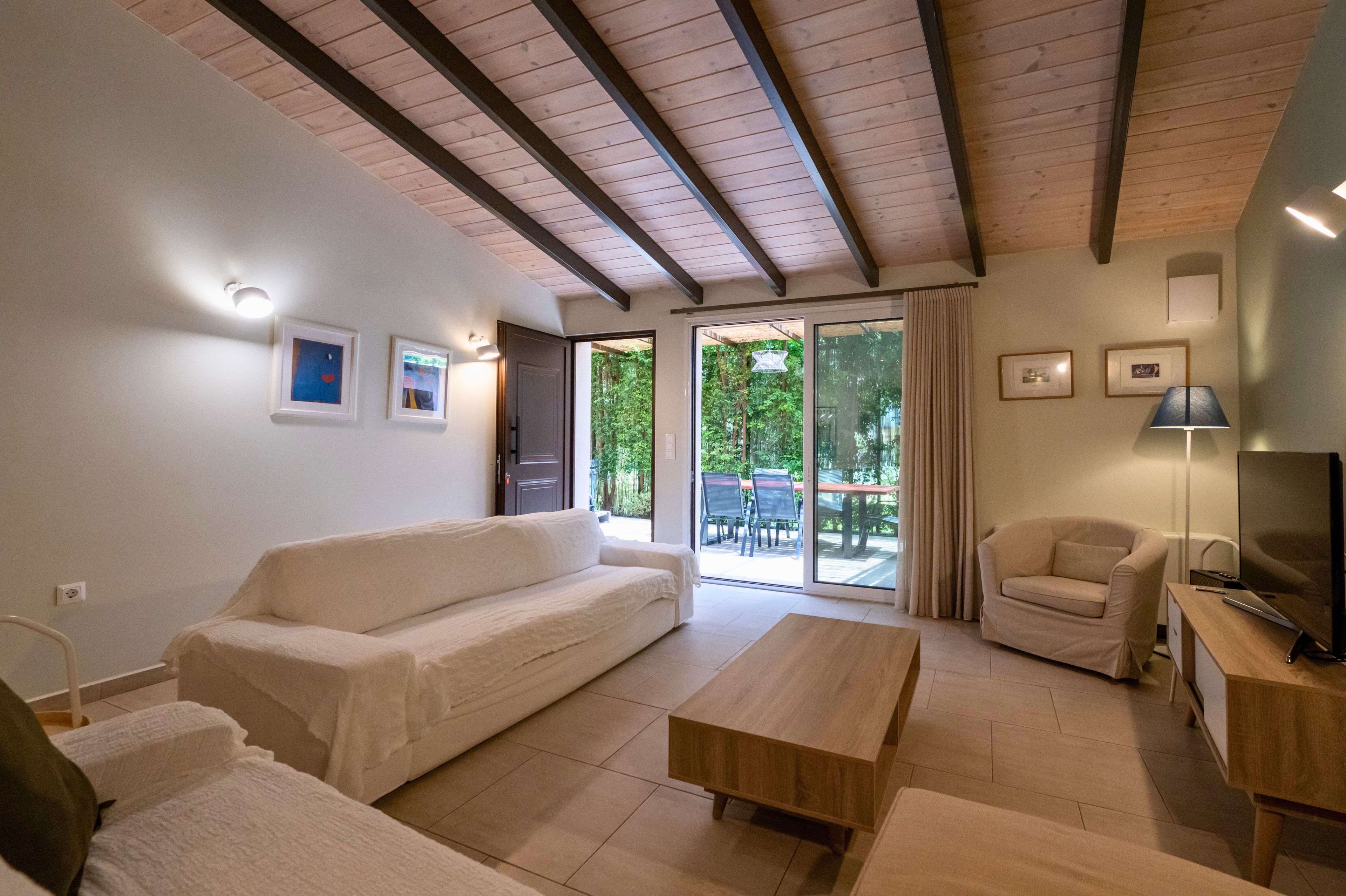 Un soggiorno moderno con soffitto in legno, un divano bianco, un tavolino da caffè in legno, un televisore su un supporto in legno e una porta scorrevole in vetro che dà su un’area esterna.