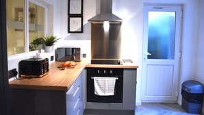 A compact kitchen with a toaster, a microwave, a stainless steel range hood, and a small oven. There is a window with a view of the outside, a potted plant, and a door with a towel hanging on it.