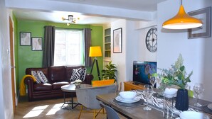 A modern living room with a green wall, a brown leather sofa, a dining table set for four, and a television.