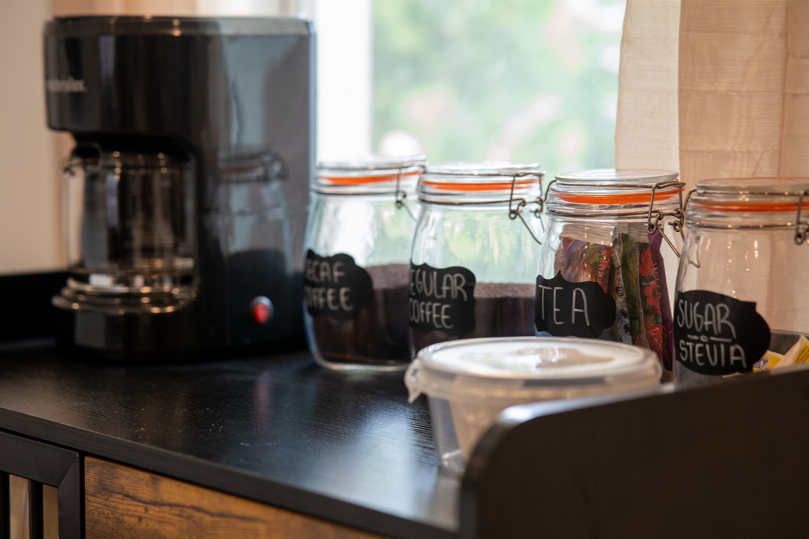 Eine Kaffeemaschine und beschriftete Gläser mit Kaffee, Tee und Zucker auf einer Küchenarbeitsplatte.