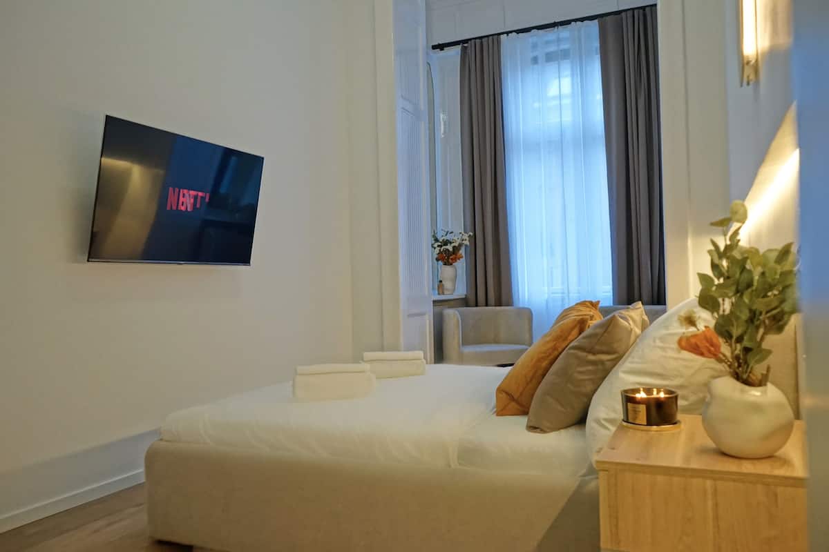 Une chambre d’hôtel avec un lit, un téléviseur fixé au mur, une table de chevet avec un vase et une bougie, et une fenêtre avec des rideaux.