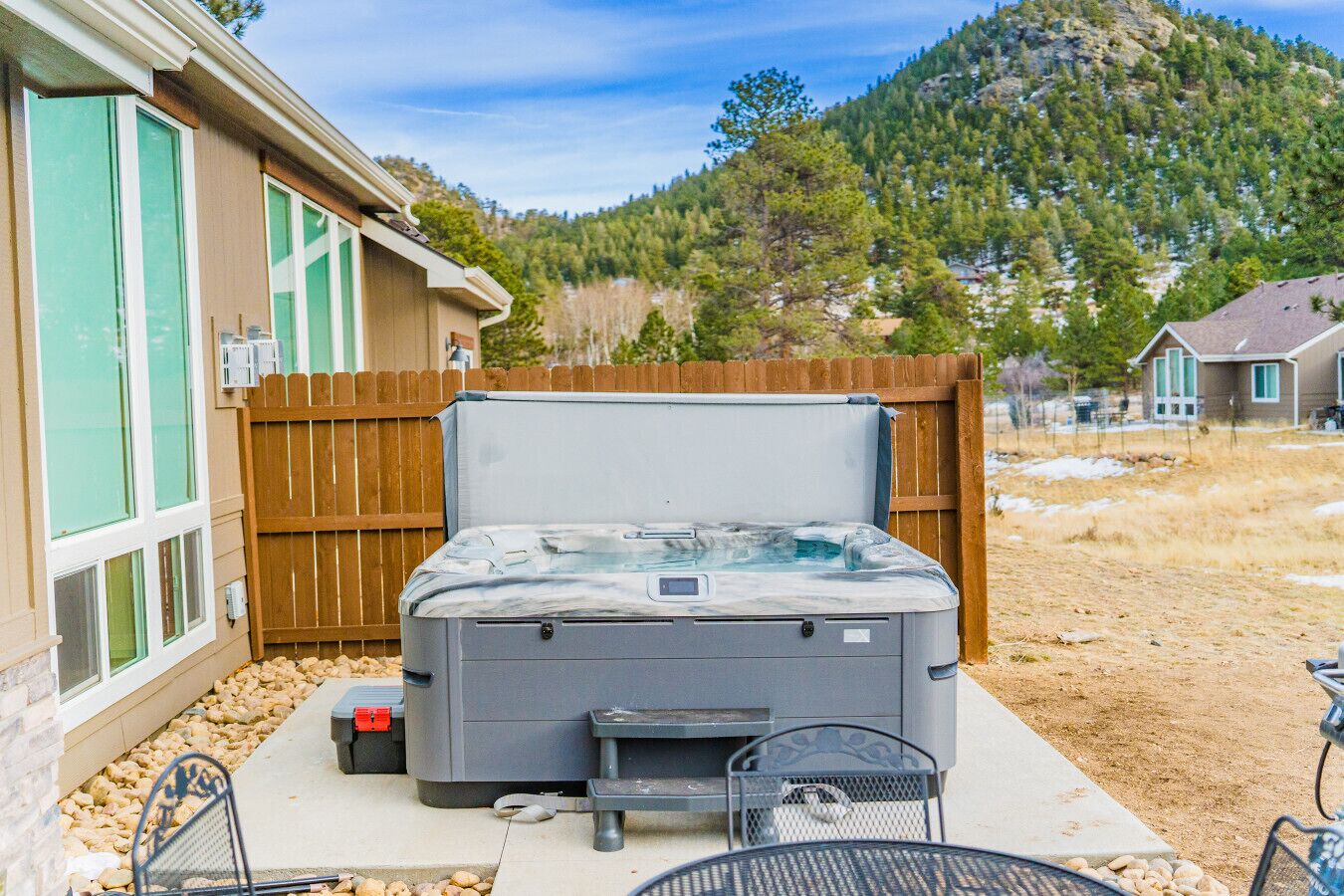 A hot tub with a wooden deck, a patio table, and chairs, surrounded by a wooden fence and a house.