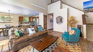 A spacious living room with a brown leather sofa, a teal armchair, and a wooden coffee table. A kitchen is visible in the background with wooden cabinets and a refrigerator. - Hot tub! Dogs OK! Views, near park! (Estes Park)