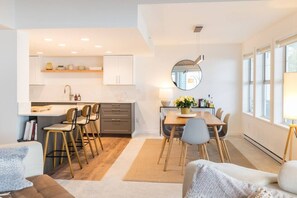 A modern kitchen with white cabinets, a dining area with a wooden table and chairs, and a large window providing natural light.