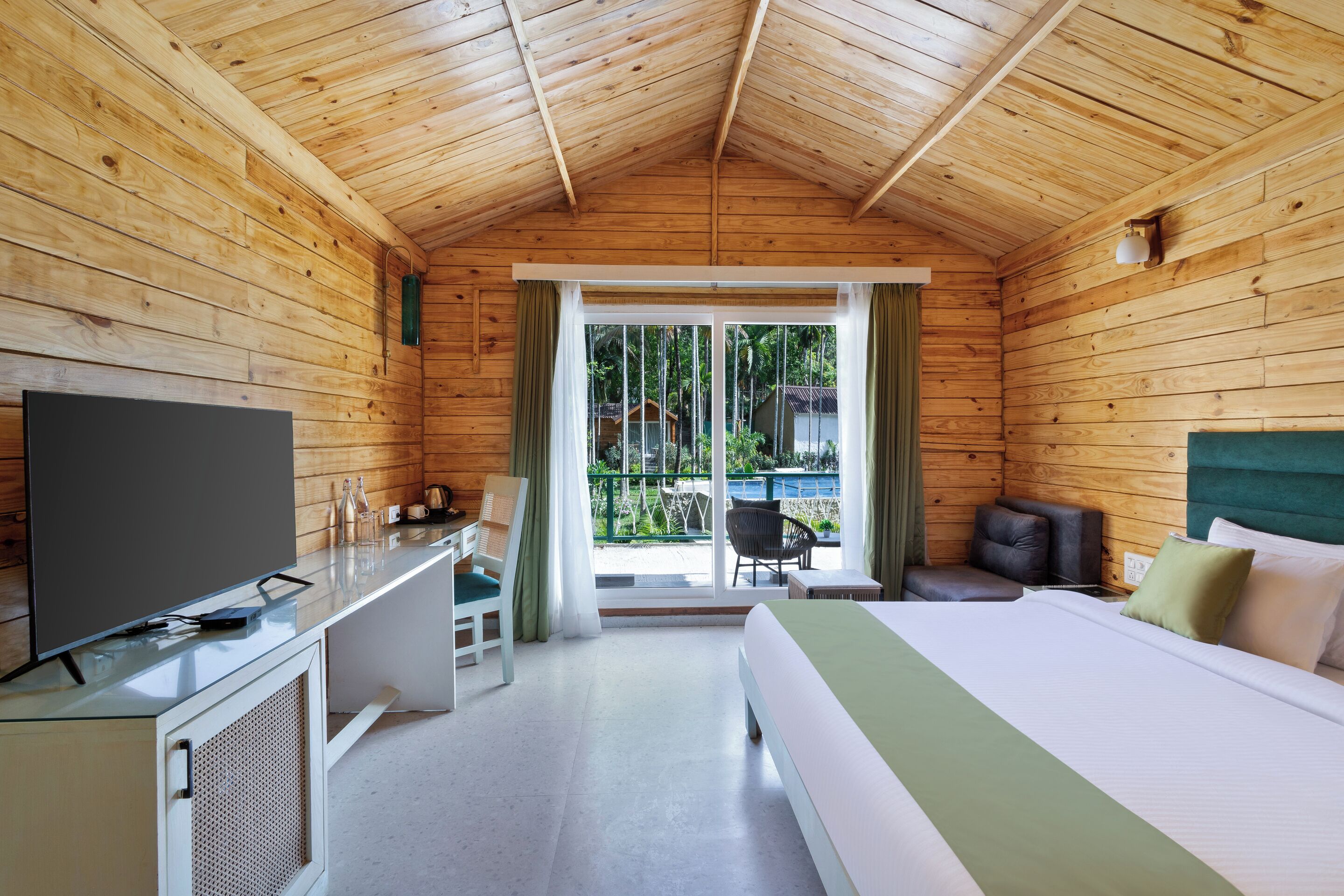 Habitación de cabaña de madera con cama, escritorio con televisor, una silla y vistas al exterior por una puerta de cristal.