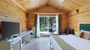 Habitación de cabaña de madera con cama, escritorio con televisor, una silla y vistas al exterior por una puerta de cristal.