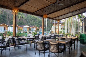 Un espacio para comer al aire libre con sillas y mesas de mimbre, rodeado de abundante vegetación y vistas a una piscina.