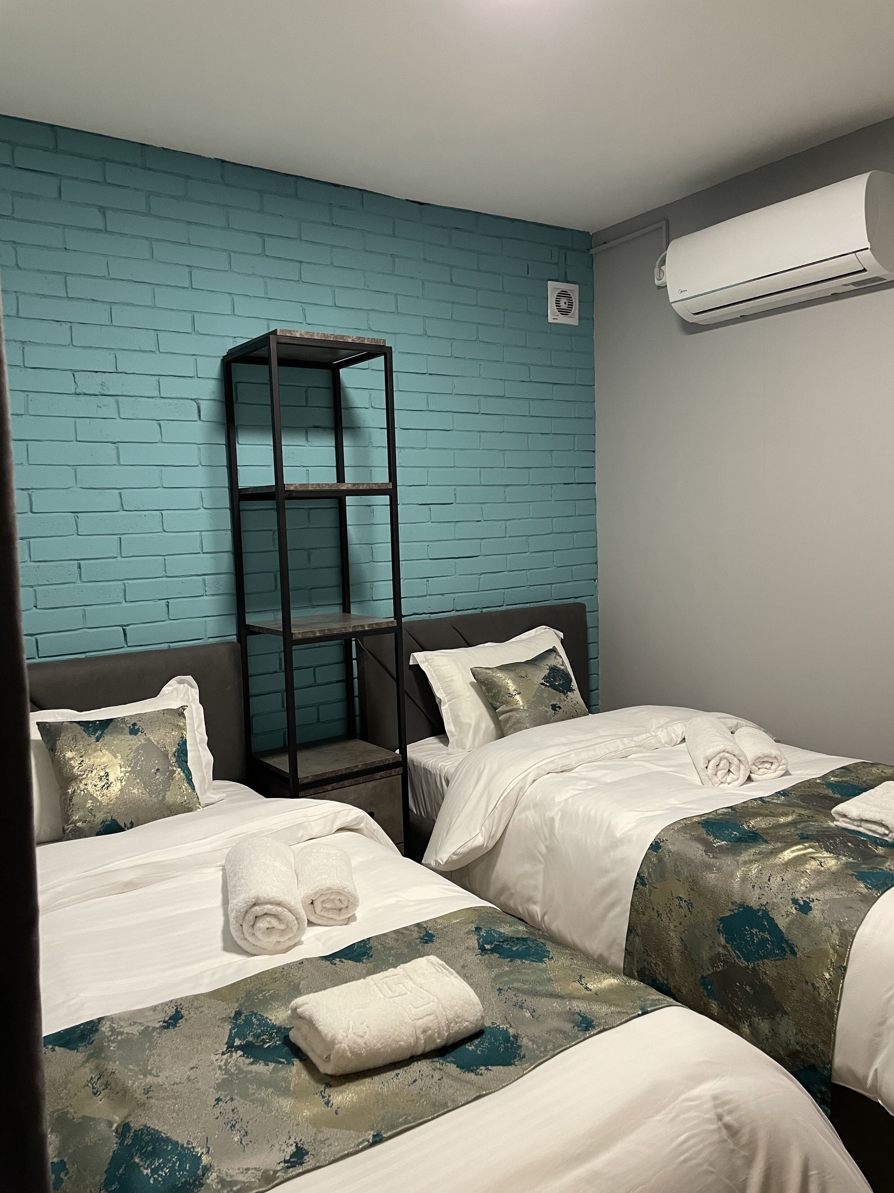 A hotel room with two beds, a metal shelf, a blue brick wall, and an air conditioning unit.