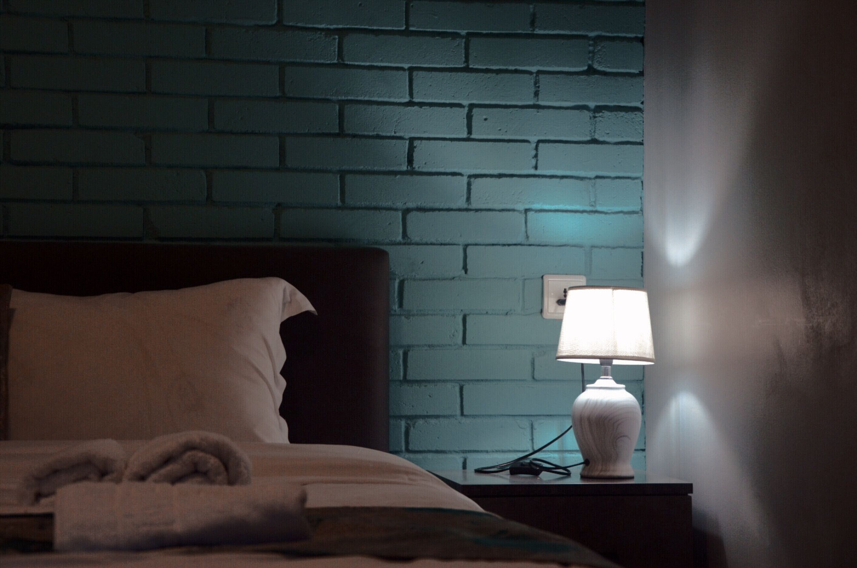 A neatly made bed with a white pillow and a folded towel, a bedside lamp on a nightstand, and a blue brick wall.