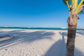 Una playa con una palmera, cielo azul despejado y barcos a lo lejos.