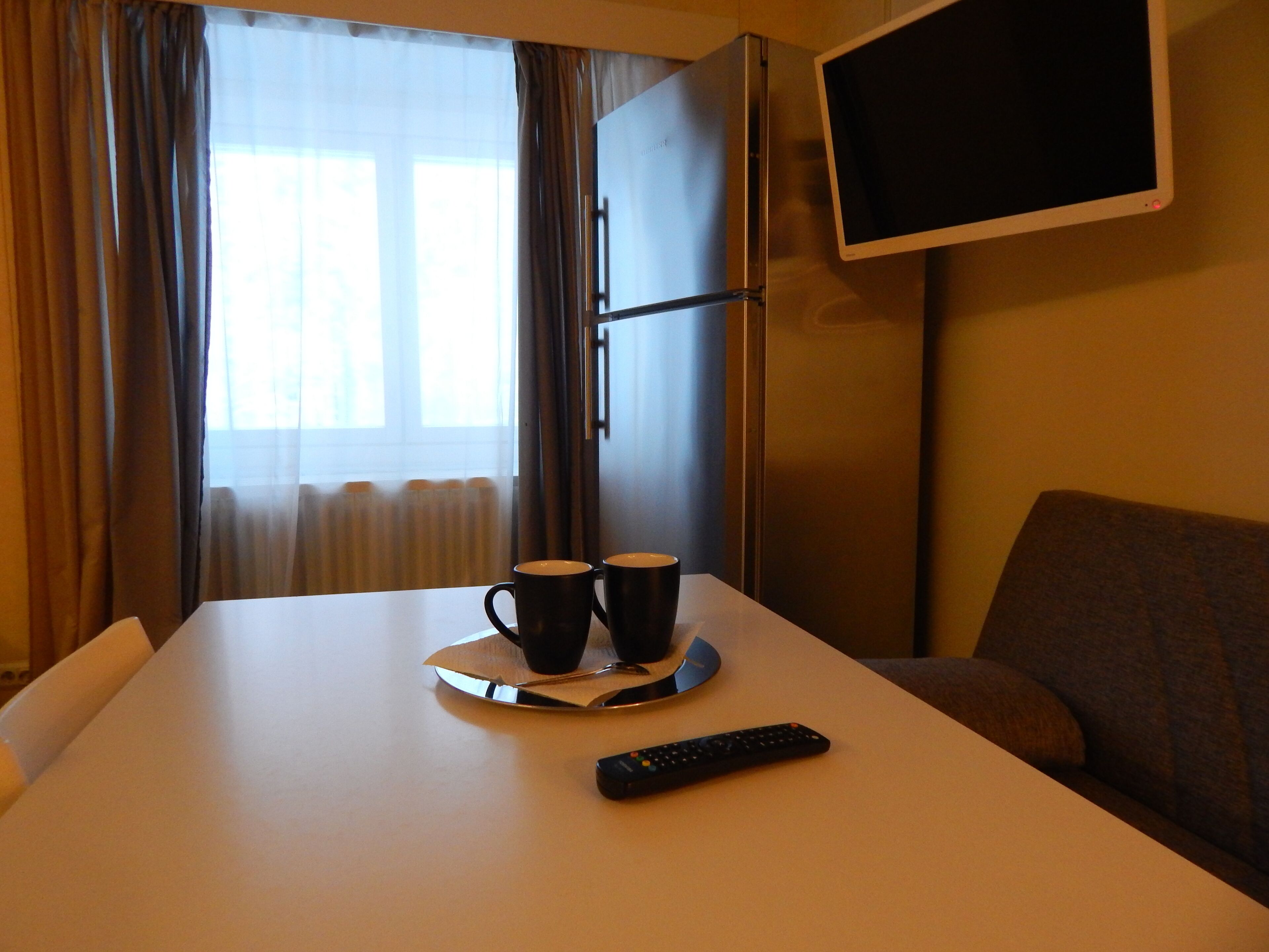 A hotel room with a bed, a TV mounted on the wall, and a table with two cups and a remote control.