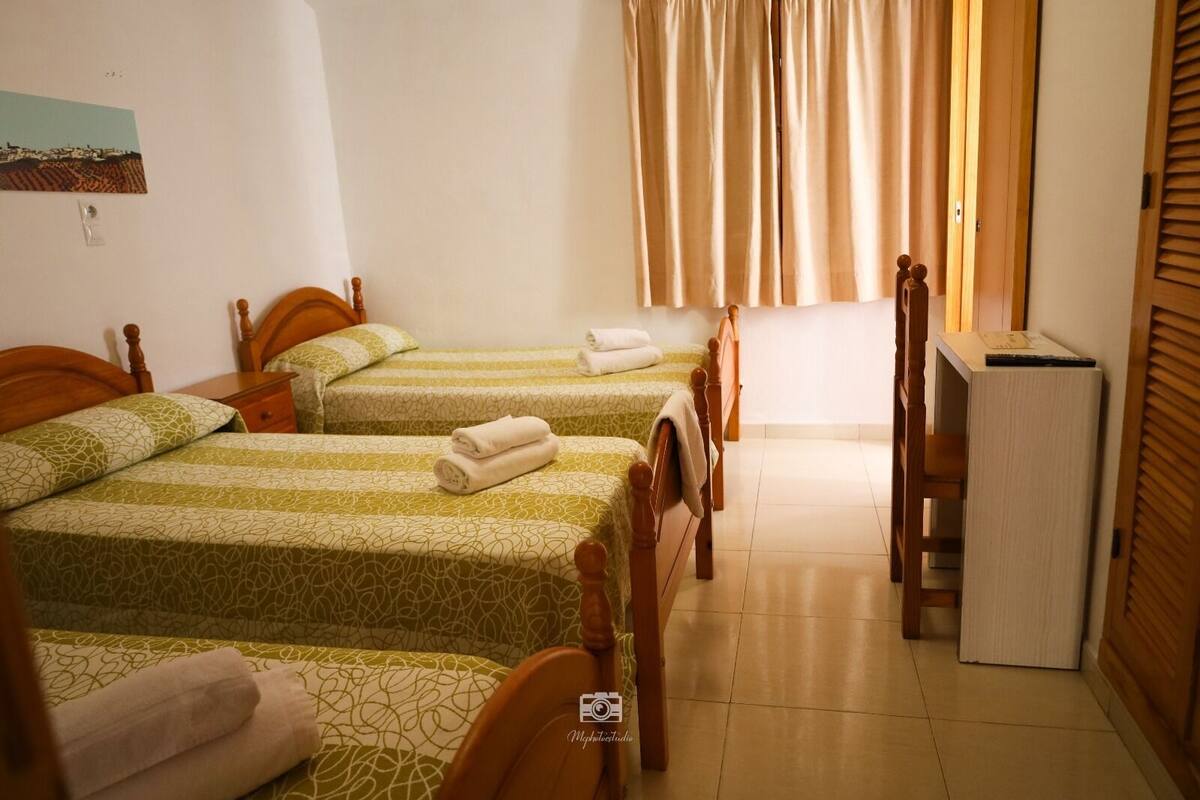 A hotel room with two single beds, a wooden wardrobe, a small desk, and a window with curtains.
