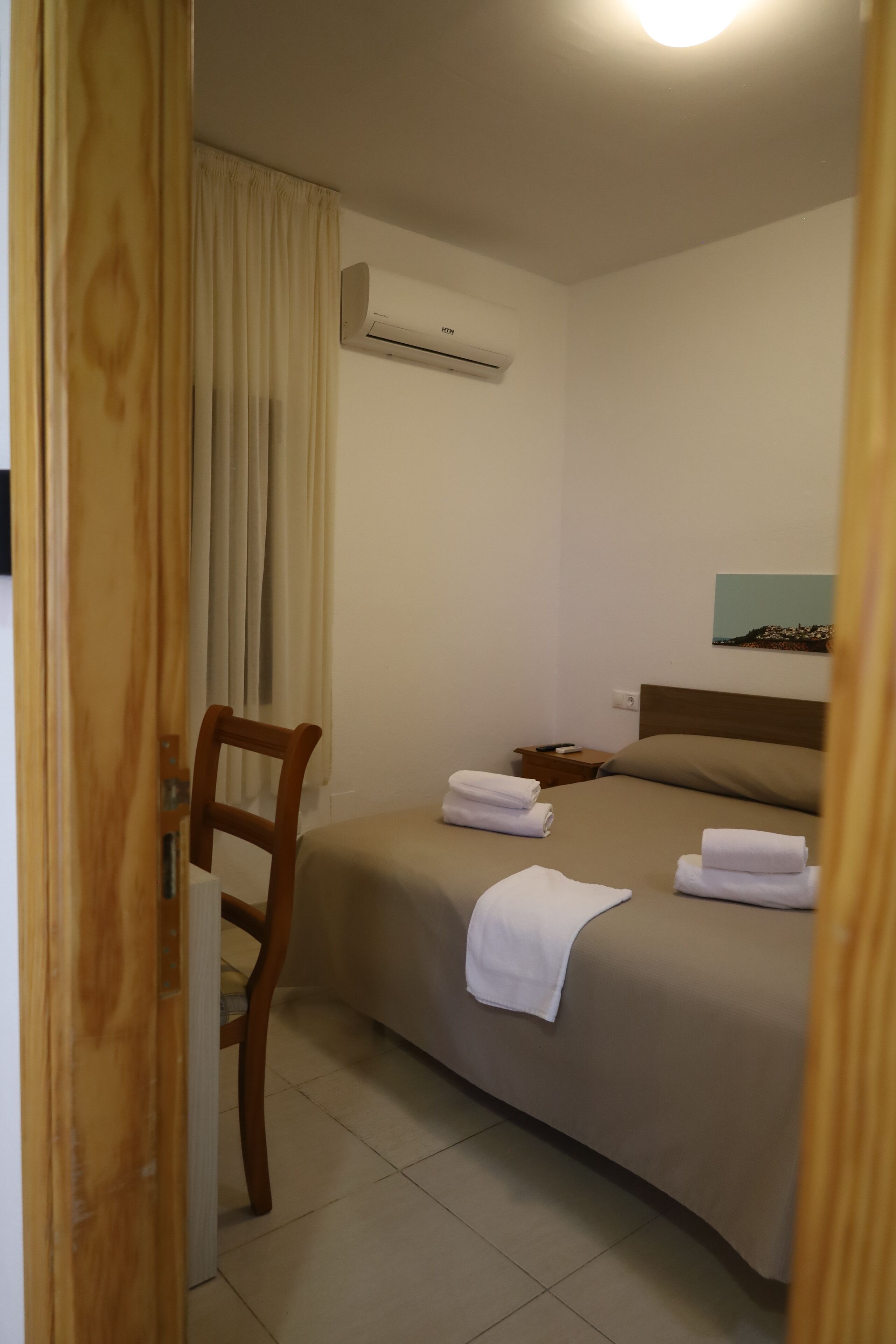 A hotel room with a bed, a chair, an air conditioning unit, and a nightstand.