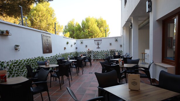 Un área de comedor al aire libre con mesas y sillas, un muro decorativo con plantas y una gran menorá.