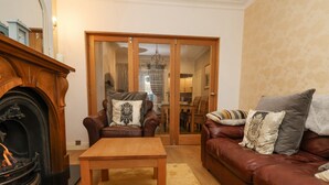 Un salon chaleureux avec une cheminée, un canapé en cuir marron, une table basse en bois et un coin repas visible à travers une porte vitrée.
