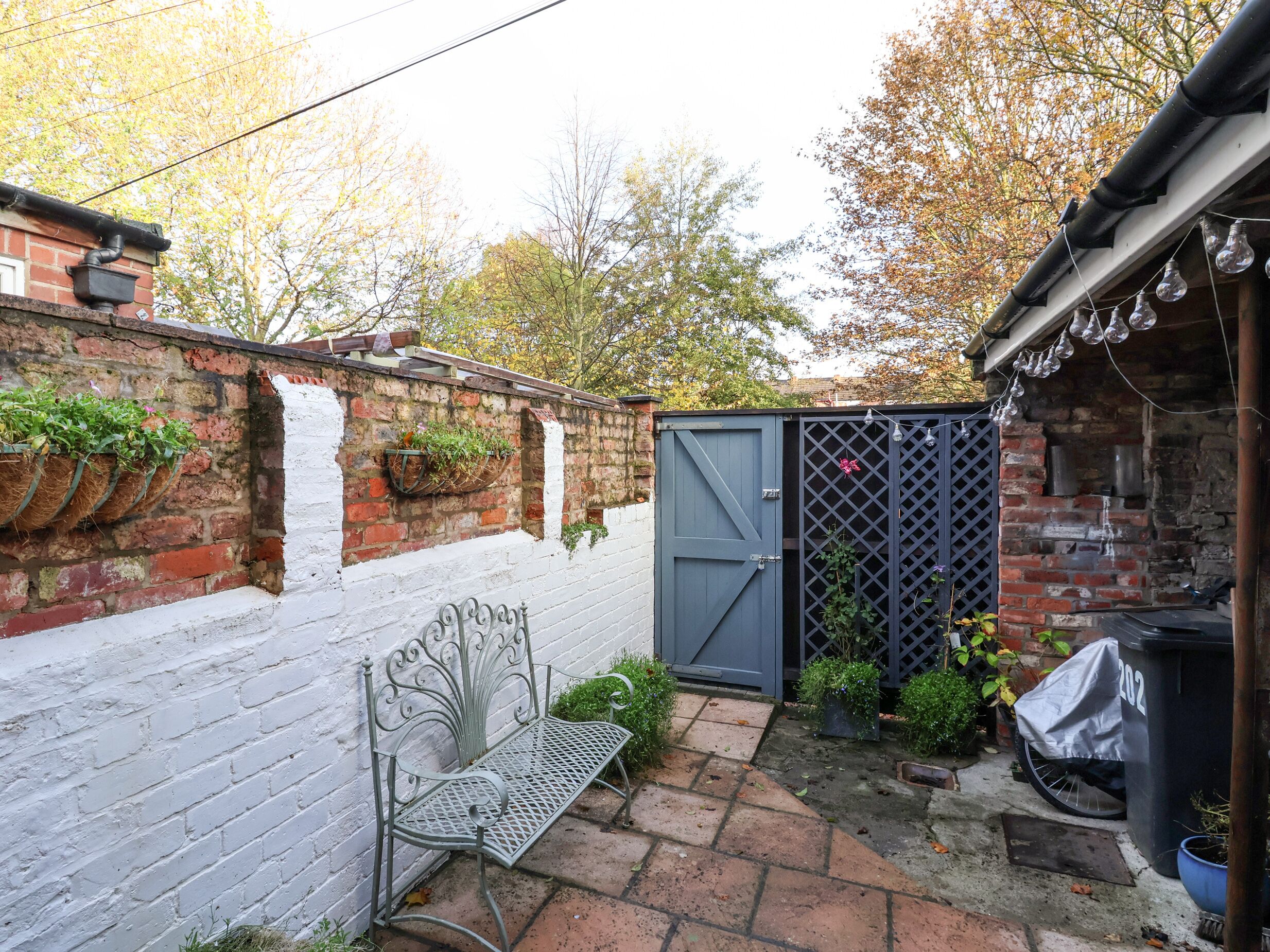 Un mur de briques orné de jardinières suspendues, un banc en métal, un portail gris et un petit espace jardin avec des plantes et un barbecue abrité.