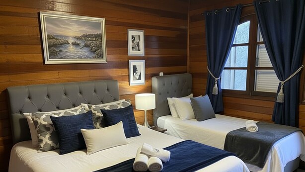 A hotel room with two beds, a large window with curtains, wooden paneling, and a painting on the wall. - Recanto Monte Suíço (Campos do Jordão)