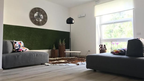 Un salon moderne avec un canapé gris, une table basse ronde, un mur d’accent vert, un lampadaire et une fenêtre donnant sur un paysage verdoyant.