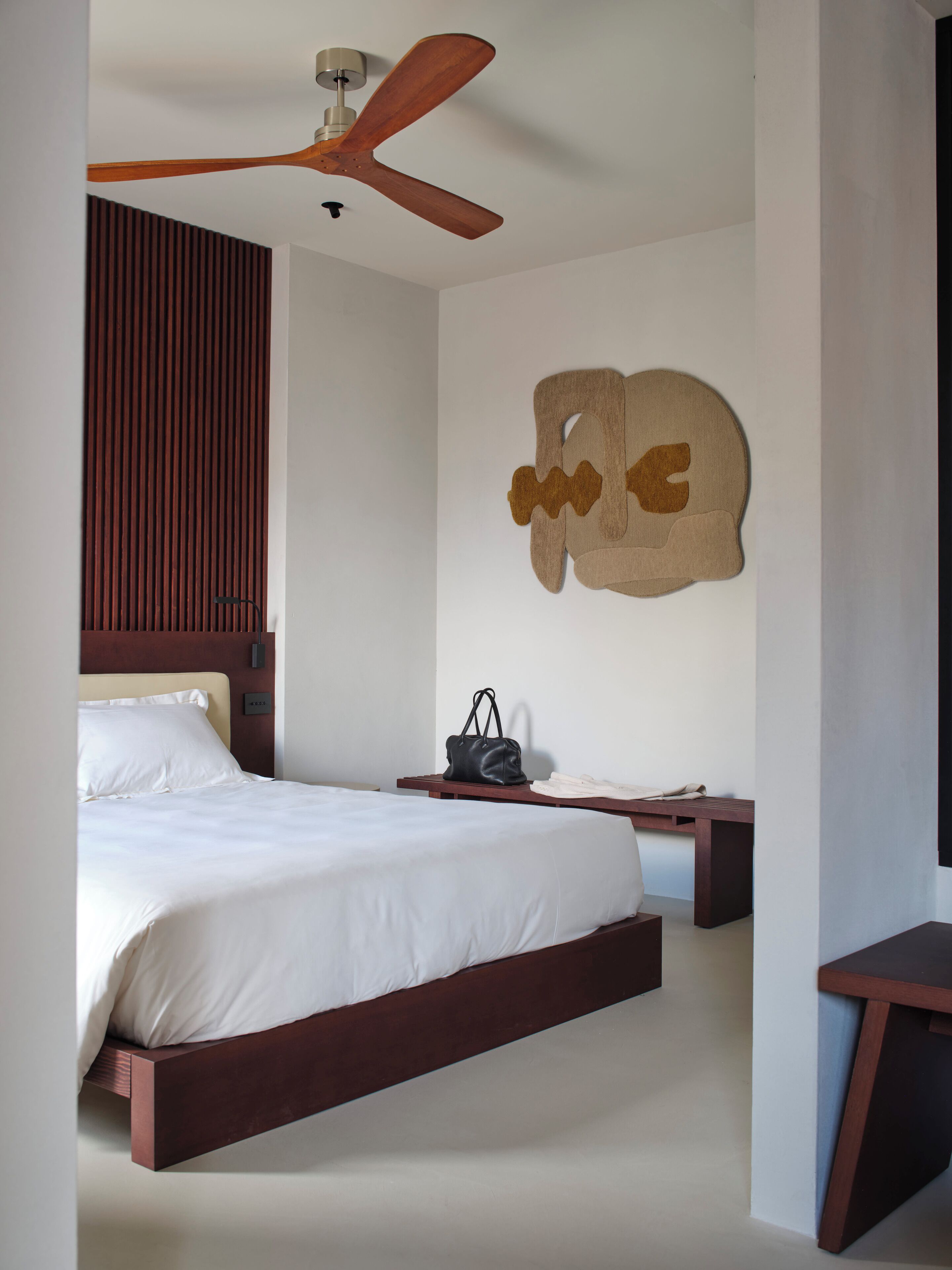 Une chambre moderne avec un cadre de lit en bois, un ventilateur de plafond, une œuvre d’art abstraite accrochée au mur et une table de chevet sur laquelle se trouve un sac à main.