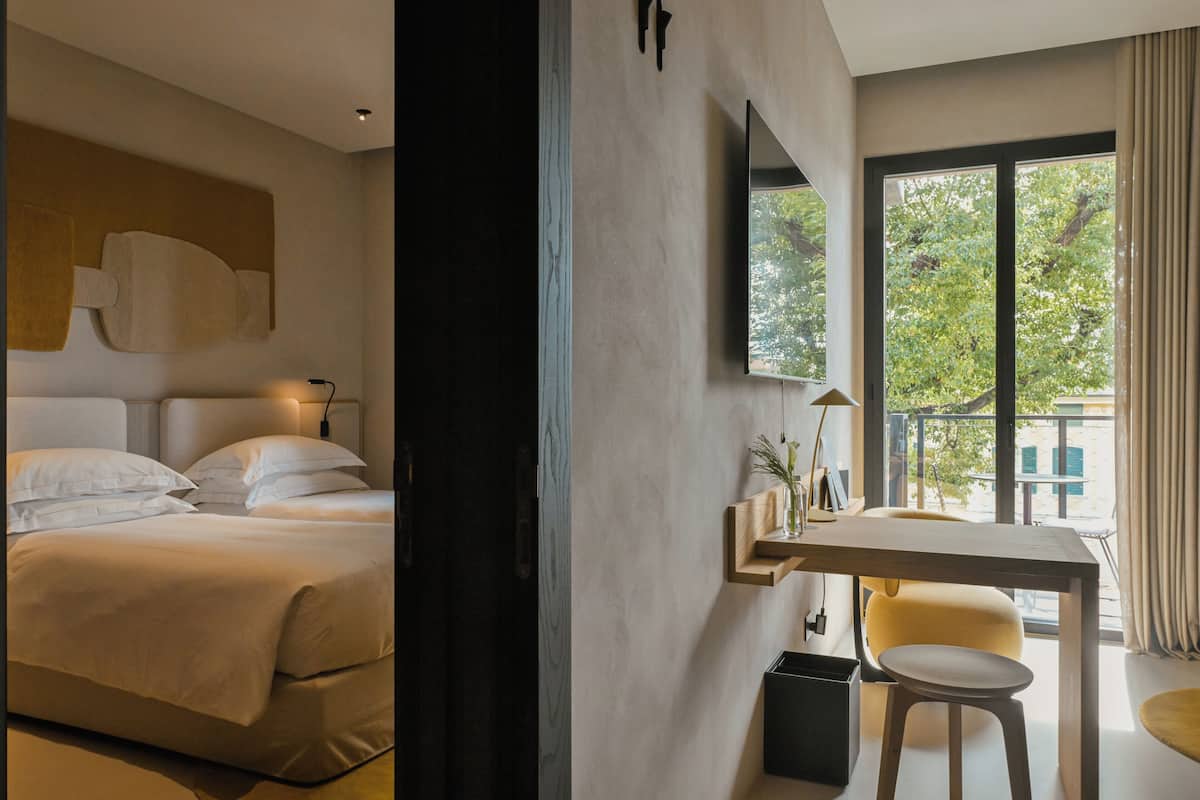 A modern hotel room with a bed, a desk, and a large window with a view of greenery.