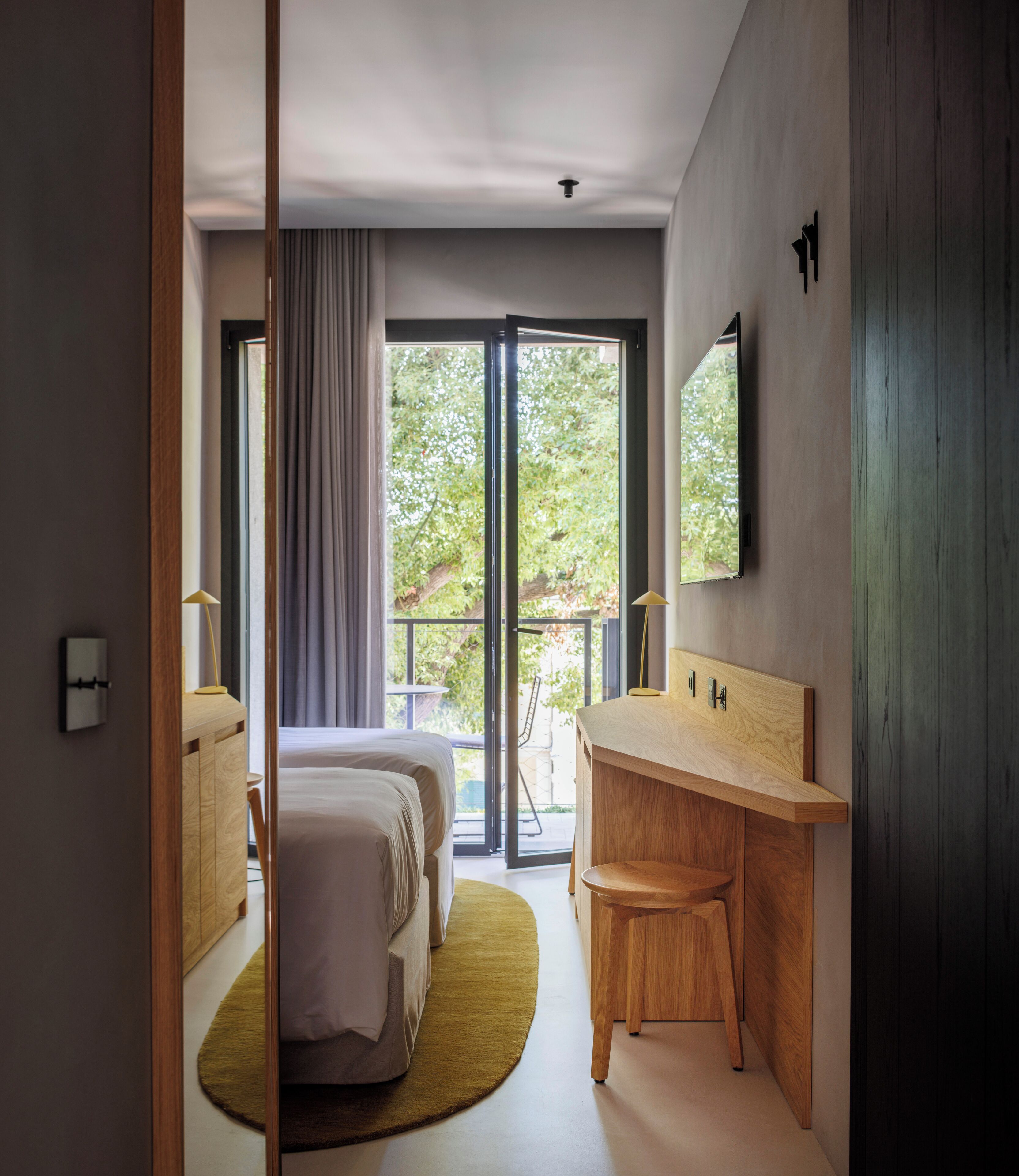 A modern hotel room with a large bed, a wooden desk, and a sliding glass door leading to an outdoor balcony.