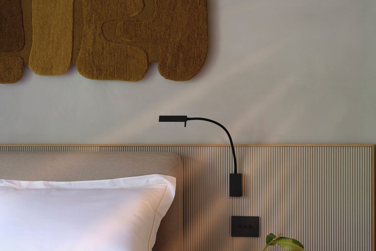 A neatly made bed with white linens, a wall-mounted lamp, and a small wooden table with a vase and a plant.
