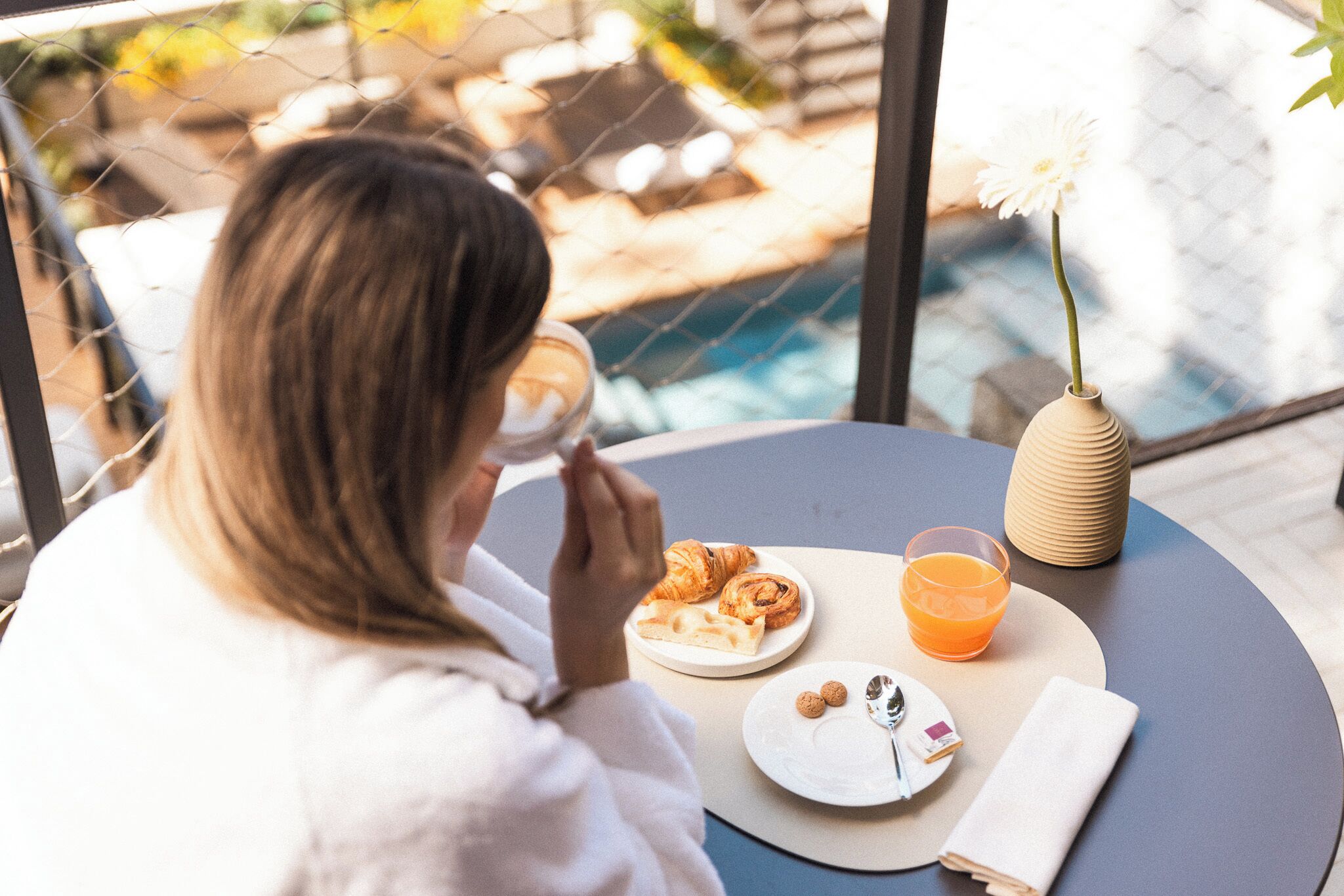 Une personne est assise à une table ronde devant laquelle se trouvent des croissants, un verre de jus d’orange et un petit vase avec une fleur.