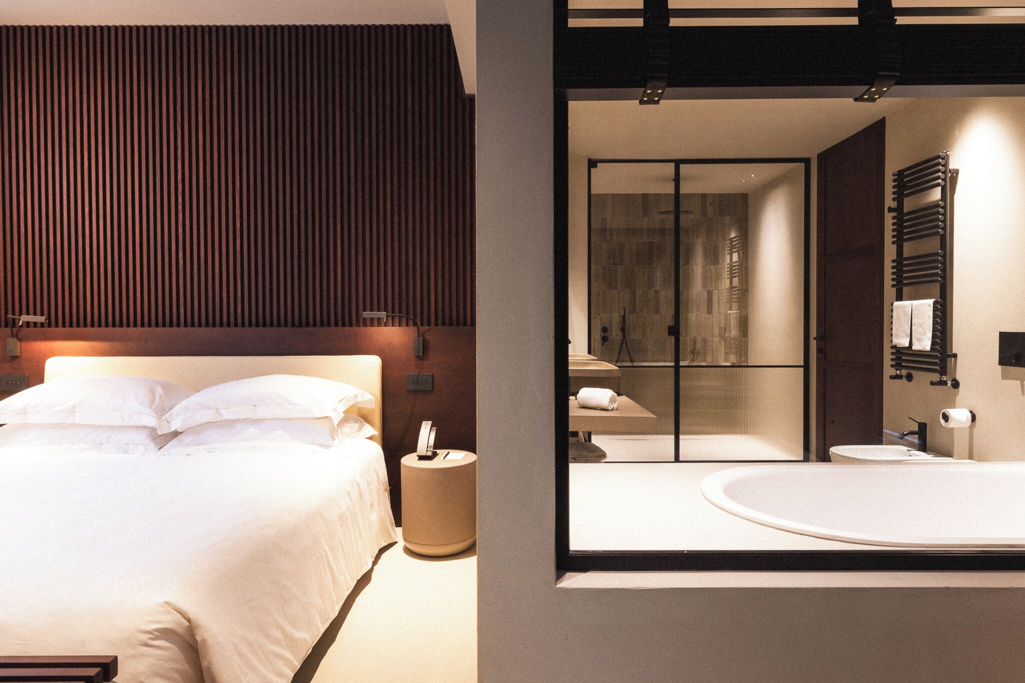 Ein modernes Hotelzimmer mit einem großen Bett, einem Nachttisch, einem runden Beistelltisch und einem Glaskastenbad mit Badewanne und Toilette.