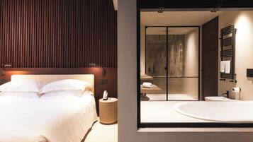 Ein modernes Hotelzimmer mit einem großen Bett, einem Nachttisch, einem runden Beistelltisch und einem Glaskastenbad mit Badewanne und Toilette.