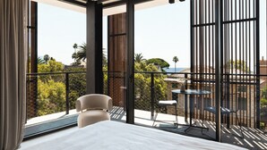 Un dormitorio moderno con una cama grande, una mesita y una silla. Tiene un balcón con vistas al mar y a la vegetación.