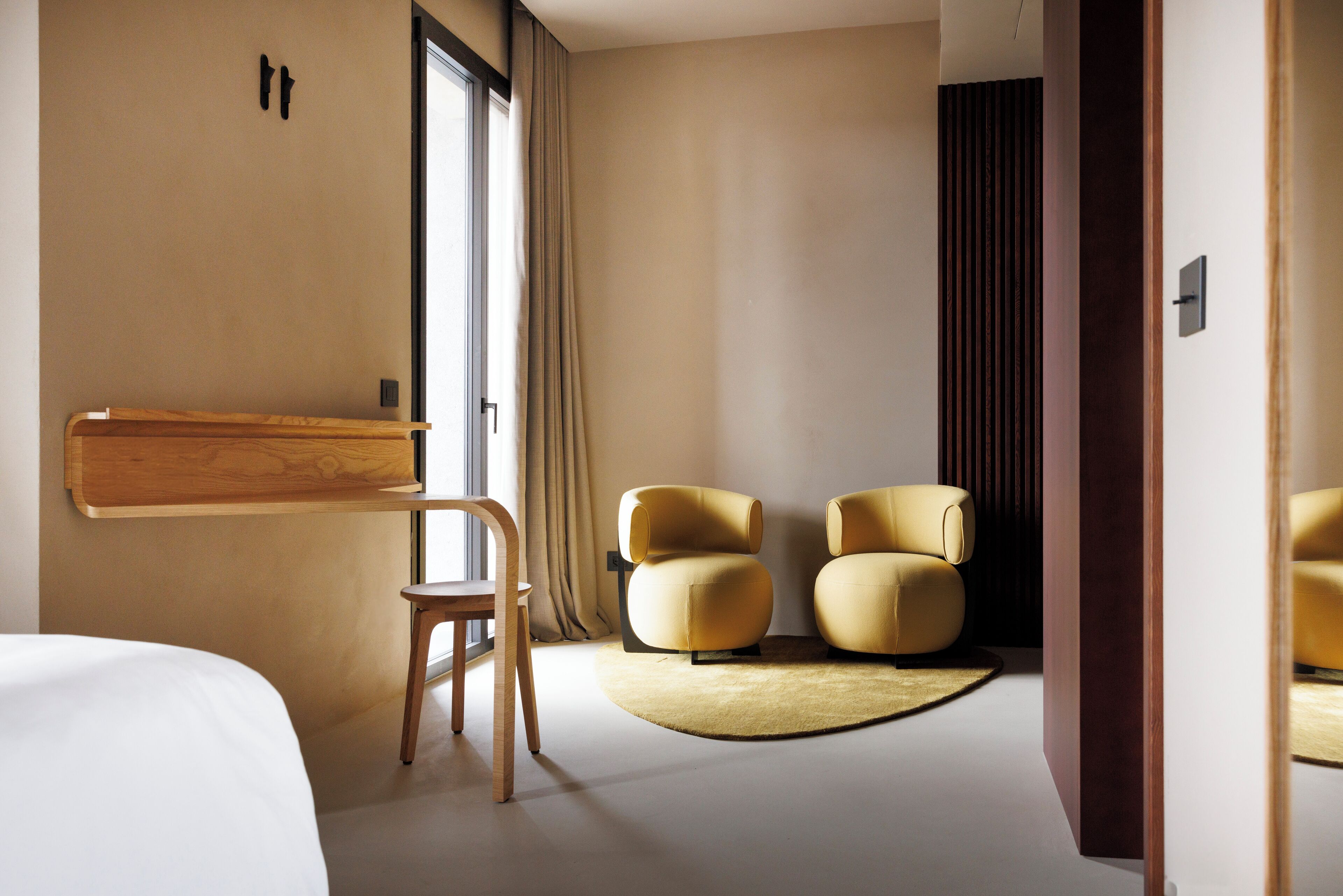 A modern hotel room with a large bed, a wooden side table, two armchairs, and a round rug.