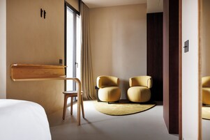 A modern hotel room with a large bed, a wooden side table, two armchairs, and a round rug.