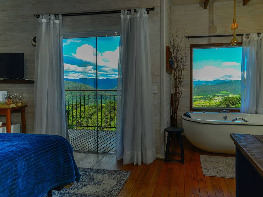 A bedroom with a large bed, a bathtub, a TV, and a balcony with a scenic view.