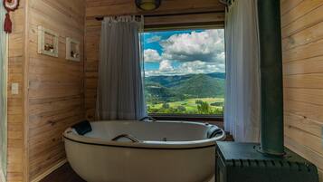 A wooden cabin room with a large bathtub, a wood-burning stove, and a window offering a mountain view.