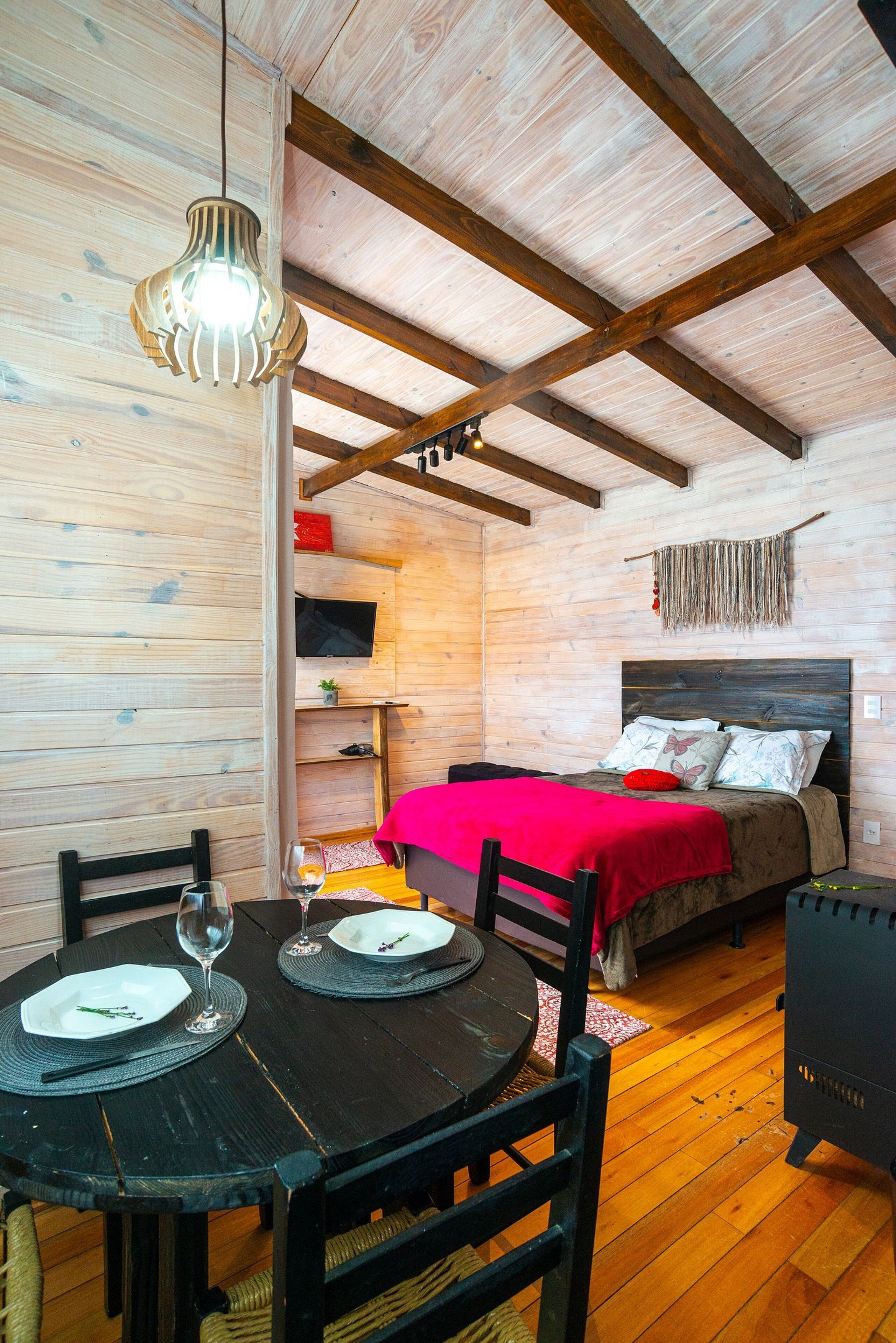 A wooden interior with a bed, a dining table with chairs, and a hanging light fixture.