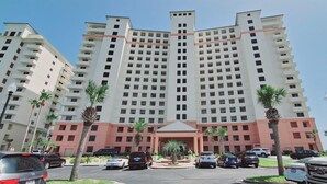 Um hotel com vários andares e entrada central, rodeado por carros estacionados e palmeiras.
