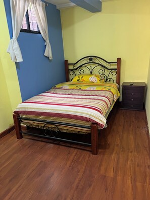 A bed with a metal frame and a colorful striped bedspread, a wooden nightstand with drawers, and a window with white curtains. - Suite Toty EN Renta (Quito)