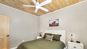 A bedroom with a bed, a nightstand, a wall clock, a ceiling fan, and a door. - Cozy 2BR/1BA cabin for 6. Fireplace, outdoor fire pit, hot tub, and soaking tub. (Norwich)