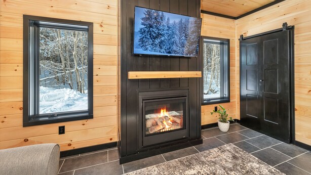 A cozy room with a fireplace, a television mounted above it, and large windows with snowy views. - Cozy 2BR/1BA cabin for 6. Fireplace, outdoor fire pit, hot tub, and soaking tub. (Norwich)