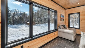 A cozy cabin interior with large windows, a sofa, and a wooden ceiling. - Cozy 2BR/1BA cabin for 6. Fireplace, outdoor fire pit, hot tub, and soaking tub. (Norwich)