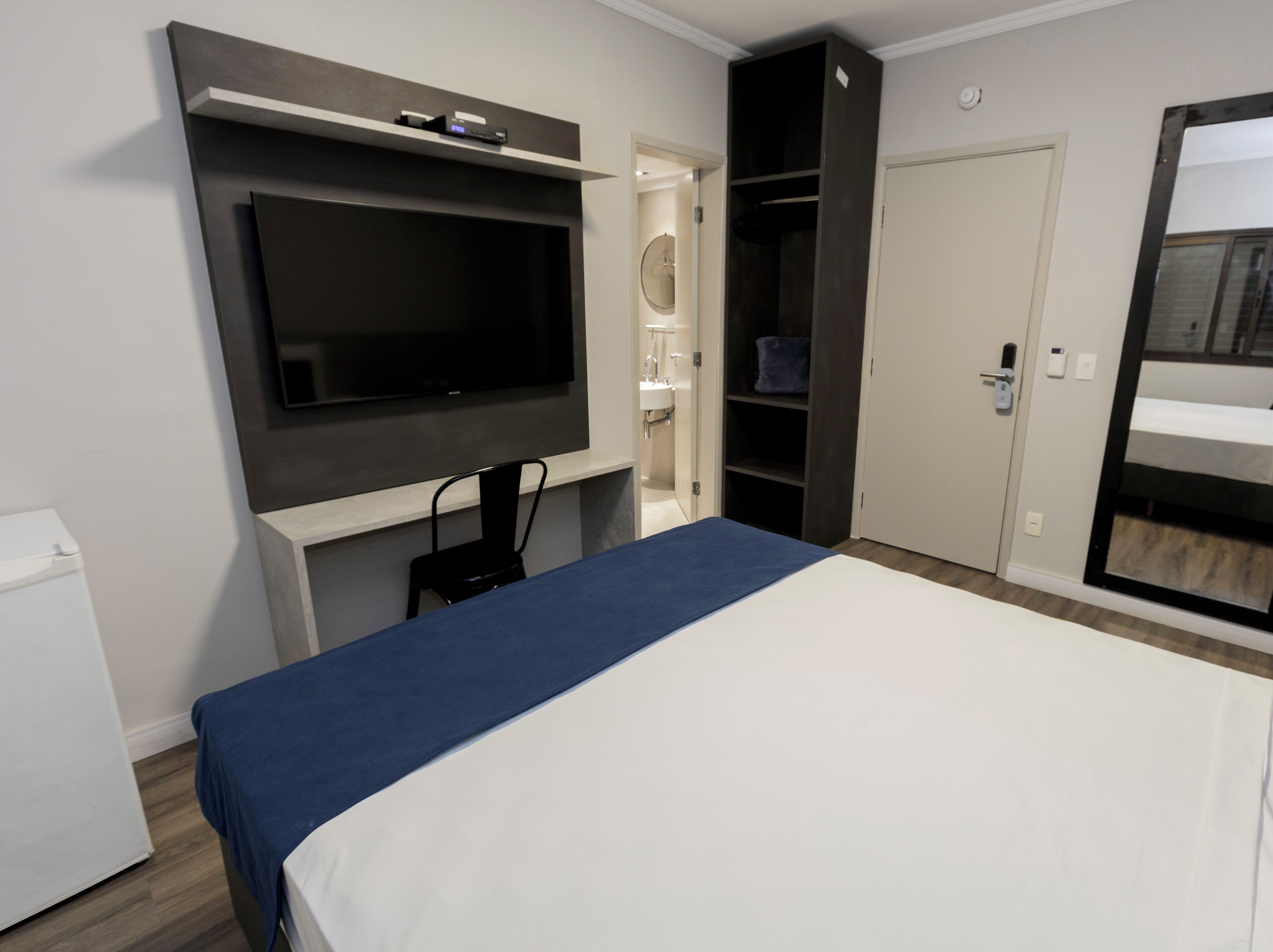 Una habitación de hotel moderna con una cama grande, un televisor de pantalla plana montado en la pared, un armario empotrado y un escritorio pequeño con una silla.