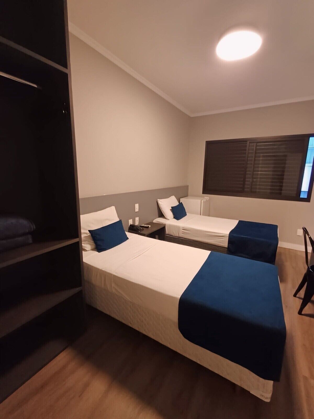 Quarto de hotel com duas camas, guarda-roupa, escrivaninha e janela com persianas.