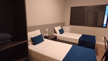 Quarto de hotel com duas camas, guarda-roupa, escrivaninha e janela com persianas.