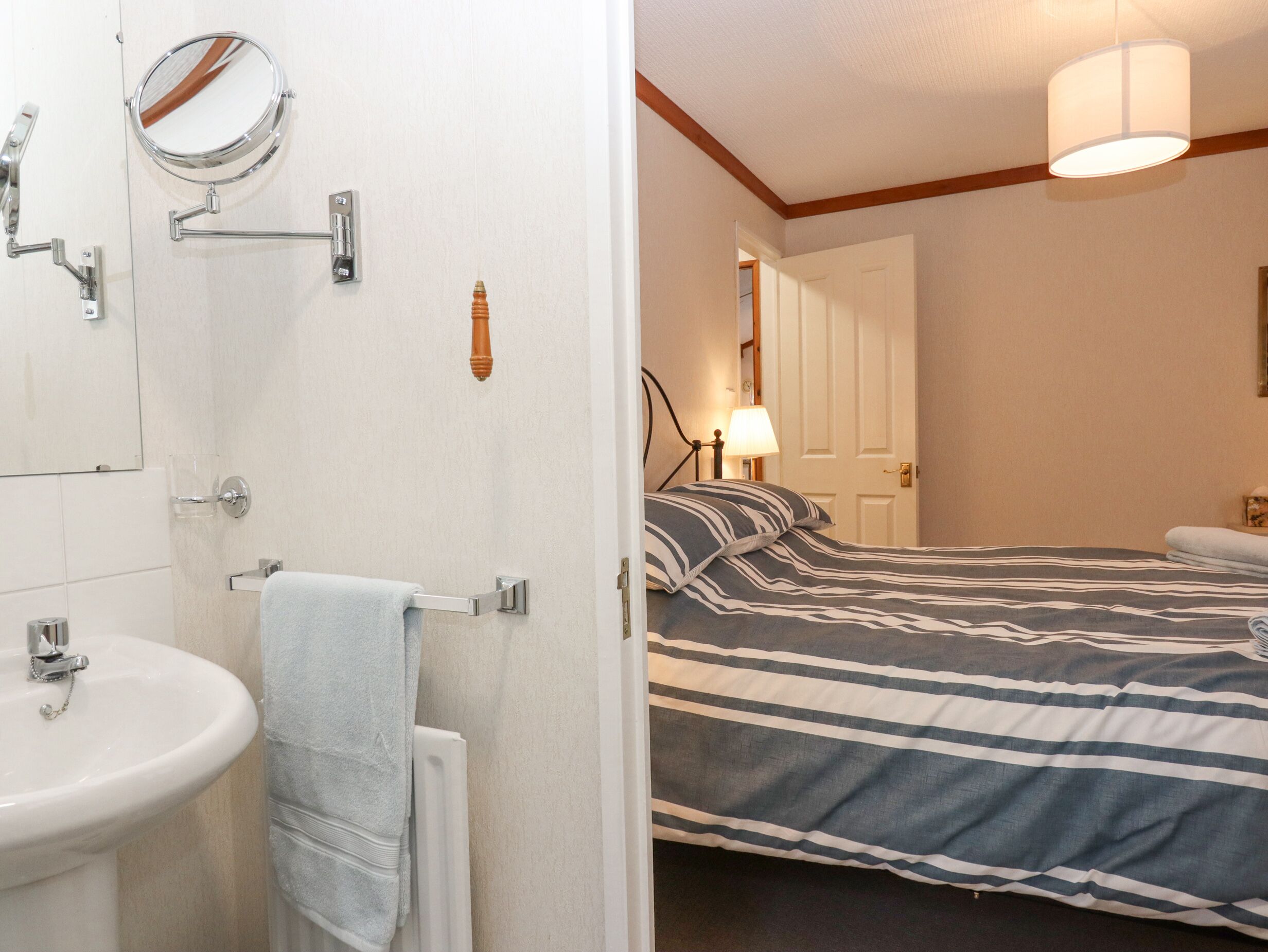 A bathroom with a sink, mirror, towel rack, and a door leading to a bedroom with a bed, lamp, and a nightstand.