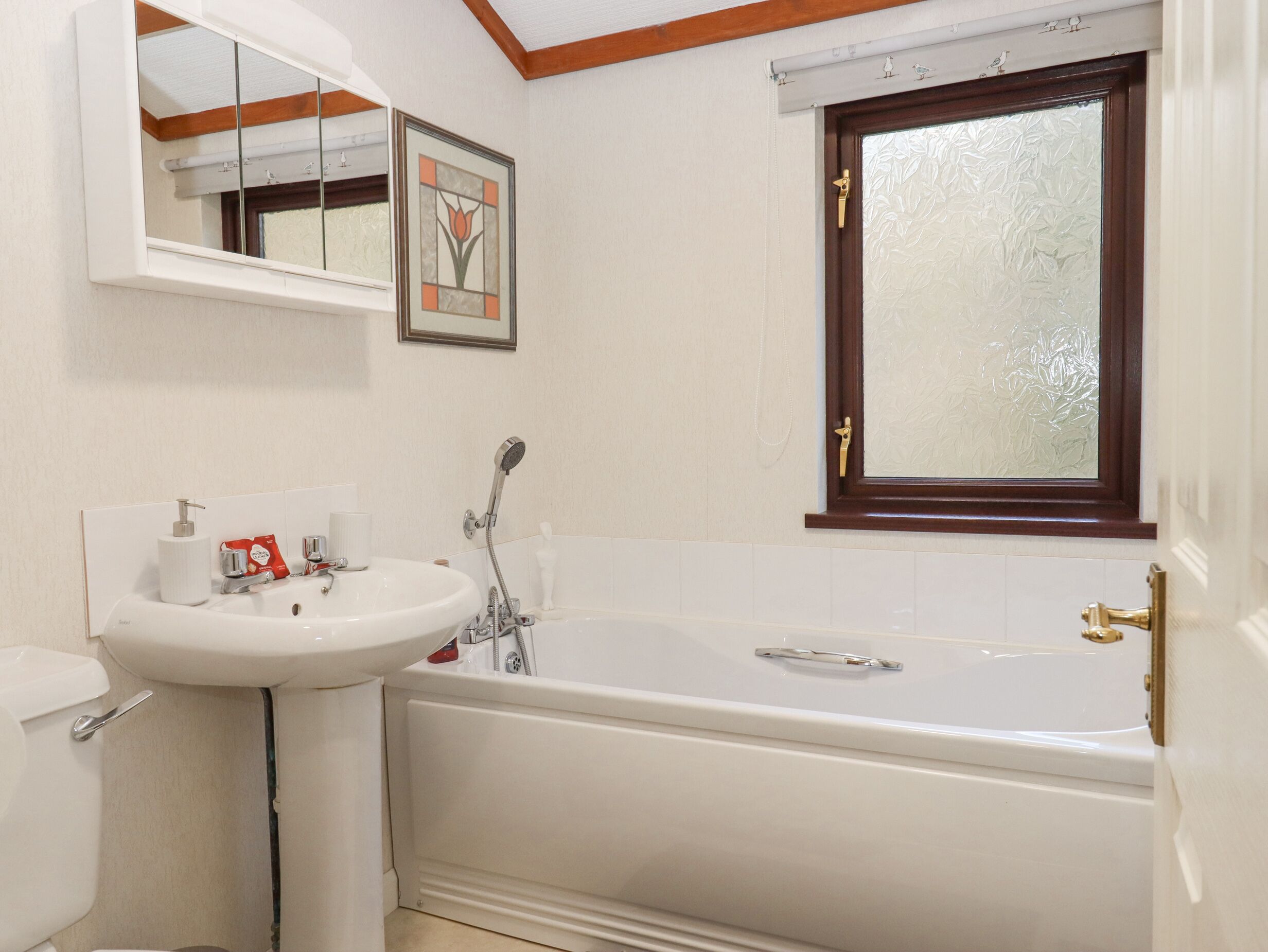 Ein Badezimmer mit weißem Waschbecken, WC und Badewanne. Ein Fenster mit Milchglas und ein gerahmtes Bild sind an der Wand angebracht.
