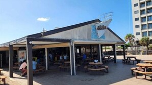An outdoor seating area with wooden tables and benches, a covered section with a sail design, and a building with a sign that reads "The Cutter." - A Slice of Heaven (Myrtle Beach)