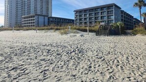 A beachfront hotel with multiple floors, a sandy beach in the foreground, and a palm tree to the right. - A Slice of Heaven (Myrtle Beach)