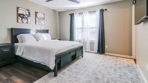 Ein Schlafzimmer mit Bett, Nachttischen, Deckenventilator, einem Fenster mit Vorhängen und einem an der Wand montierten Fernseher.