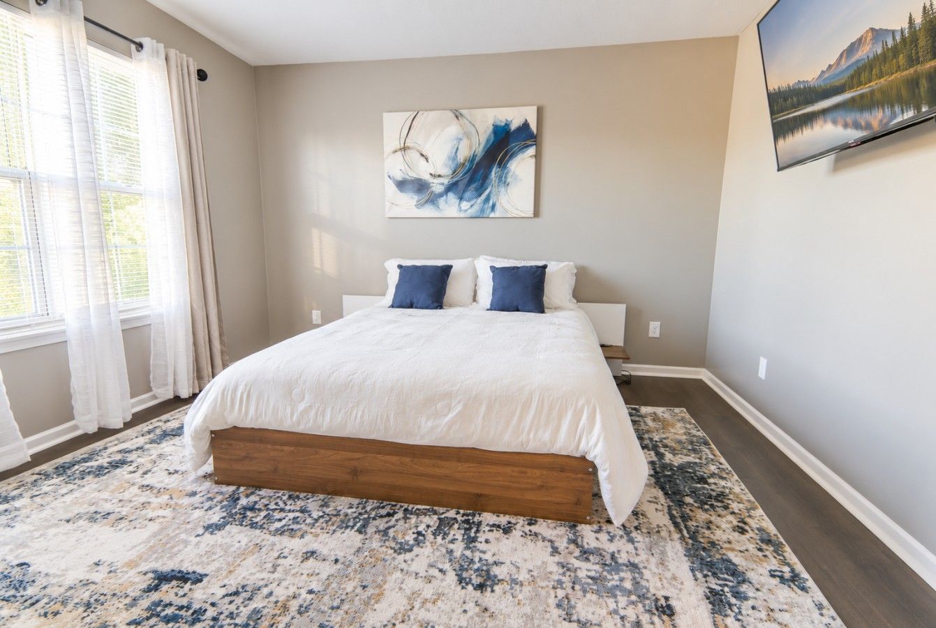 Une chambre à coucher avec un lit, une grande fenêtre avec des rideaux, un tableau au mur et un tapis.