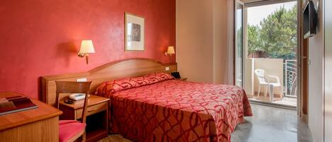 Une chambre d’hôtel comprenant un lit, un bureau avec une chaise, une table de chevet, une lampe et une porte-fenêtre coulissante donnant sur un balcon équipé d’une chaise.